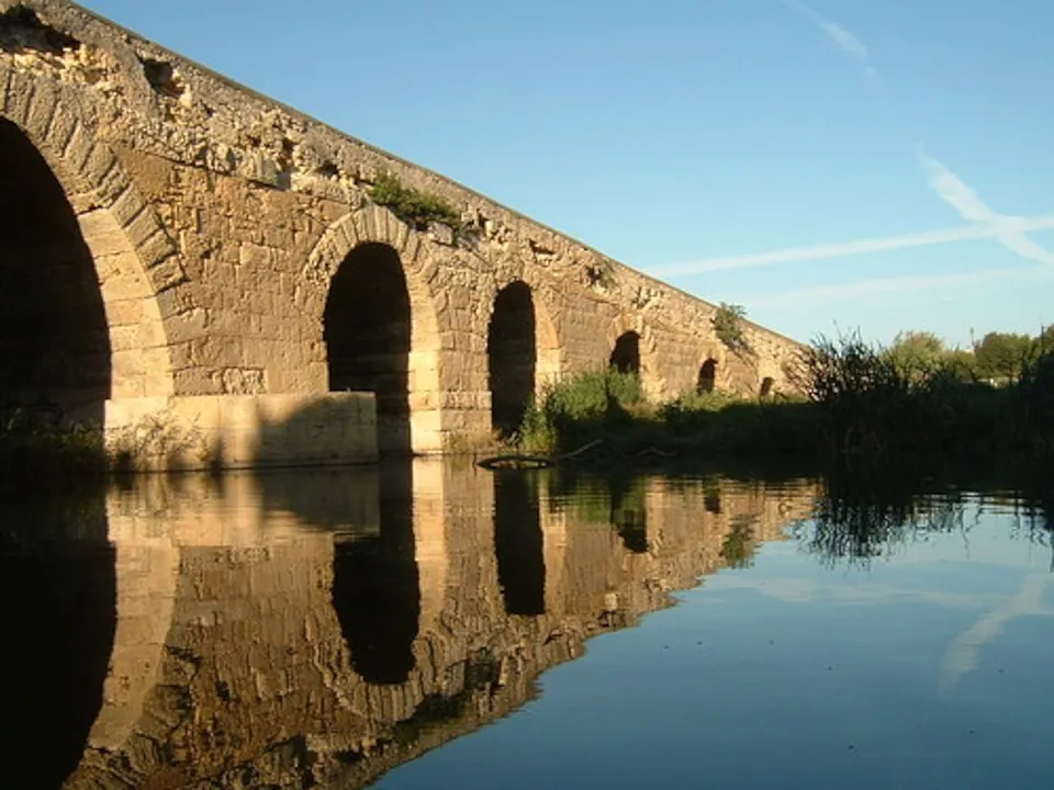 Round stone arches of a long bridge reflected in still water, with reeds growing at its far end under a blue sky.