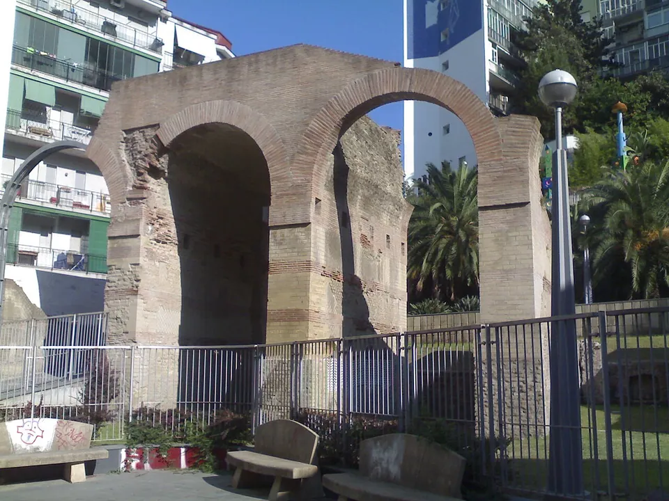Two tall round brick arches of a ruined bridge stand behind railings in a small paved park, with benches in front and apartment blocks and palm trees around it.