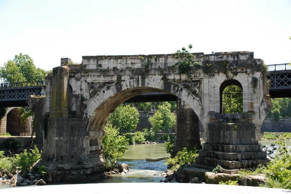 A single stone arch on two broad piers standing alone in a river, overgrown with plants along its top, with modern iron bridges and trees on the banks behind it.