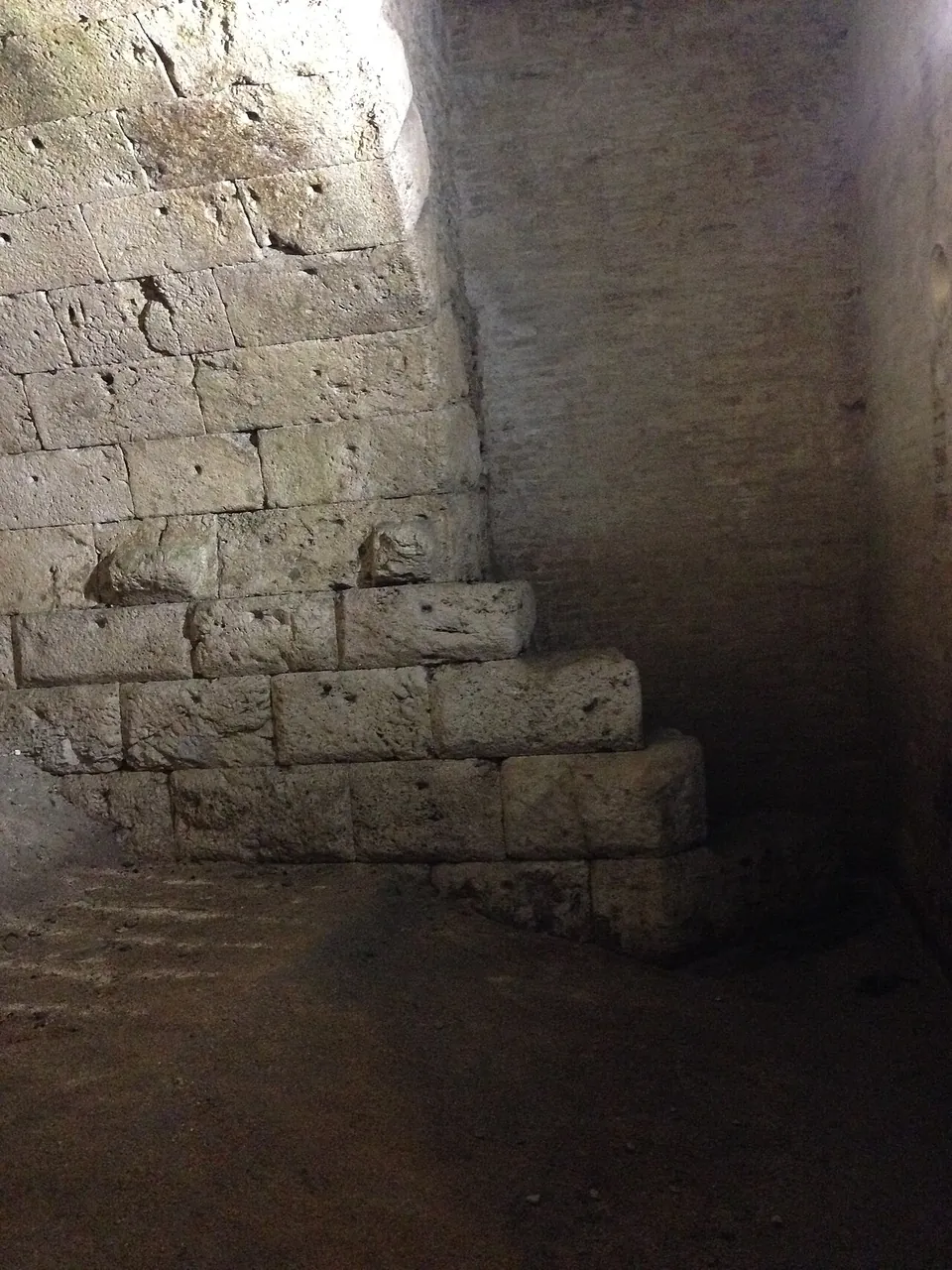 Squared stone blocks laid in steps at the foot of a wall in a dim underground chamber with an earth floor.