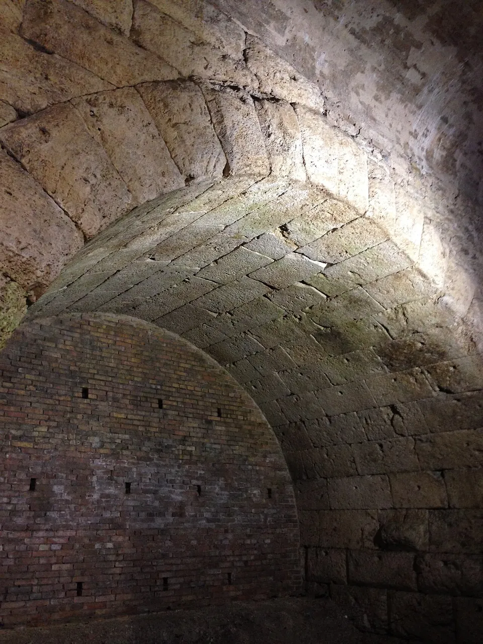 The underside of a barrel vault of squared stone blocks, lit from one side, its far end closed by a wall of brick.