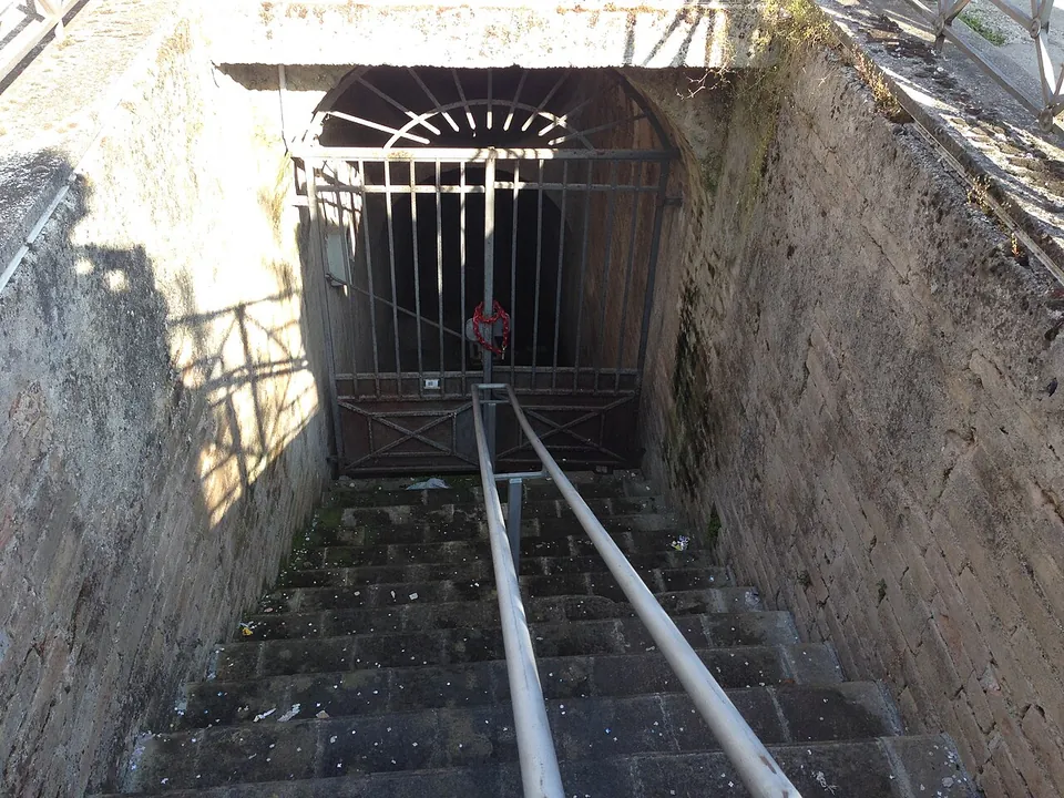 A worn stone stair with a central handrail running down between stone walls to a closed iron gate under an arched opening.