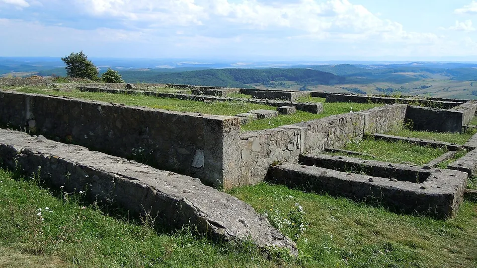 Restored stone walls, about knee to waist high, outlining rooms and a long hall on a grassy hilltop, with a wide view over wooded hills.