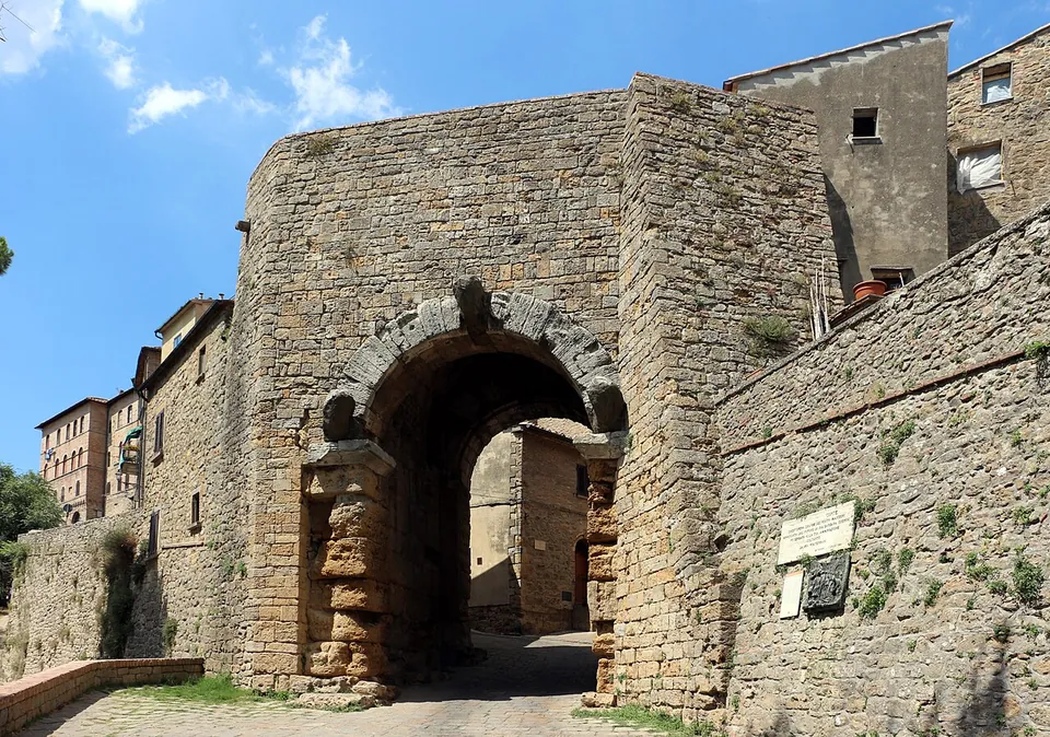 A city gate of large, roughly squared stone blocks, its round arch framed by three weathered carved heads, one at the top and one at each springing, set in a tall wall with houses behind and a plaque beside it.