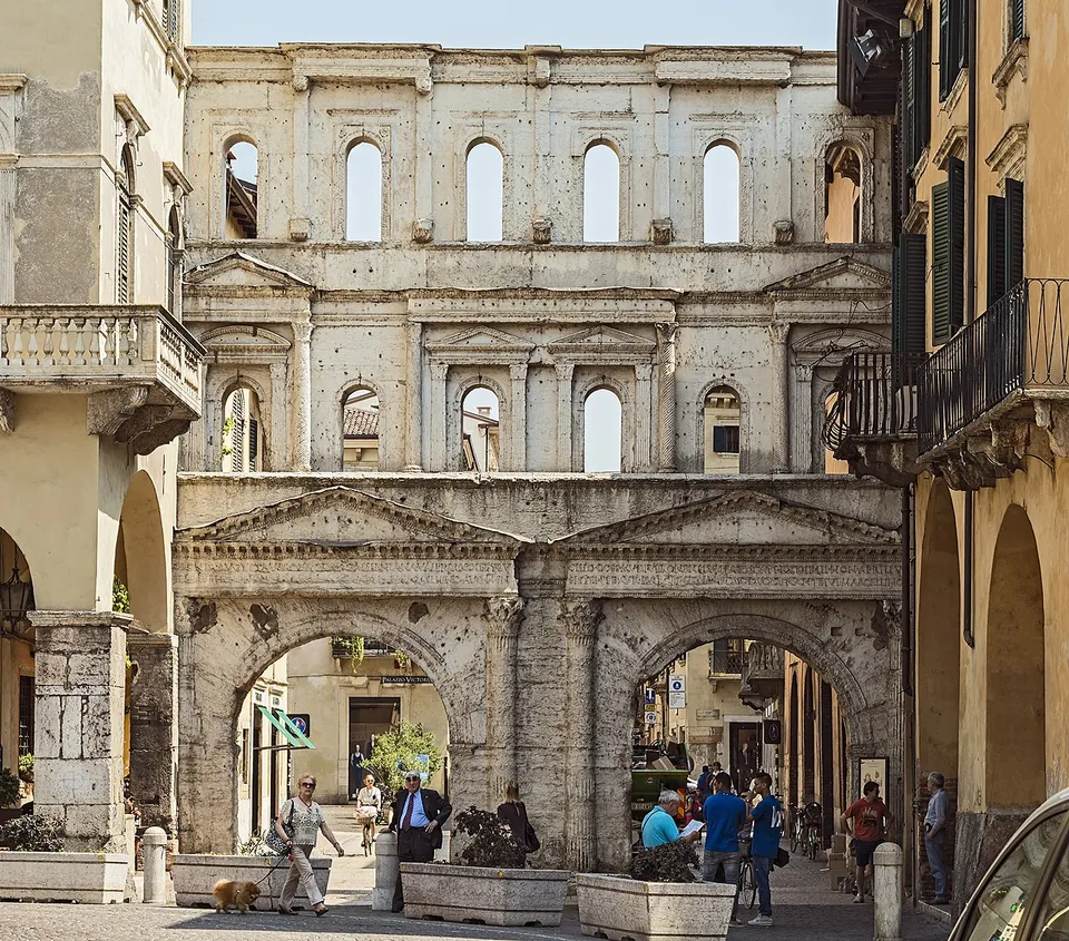 A tall pale stone facade filling the end of a street between later houses with balconies, two round arches at street level each framed by columns and a triangular pediment, and above them two storeys of arched windows open to the sky; people walk through the arches.