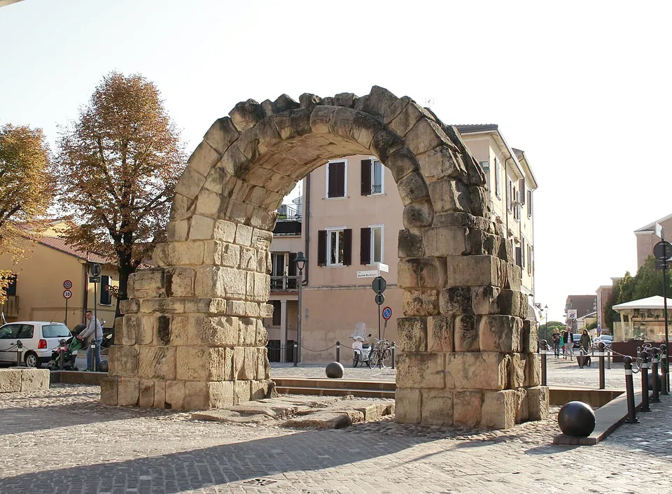 A single round arch of large rough-cut stone blocks standing on its own in a paved square, with houses and trees behind it.