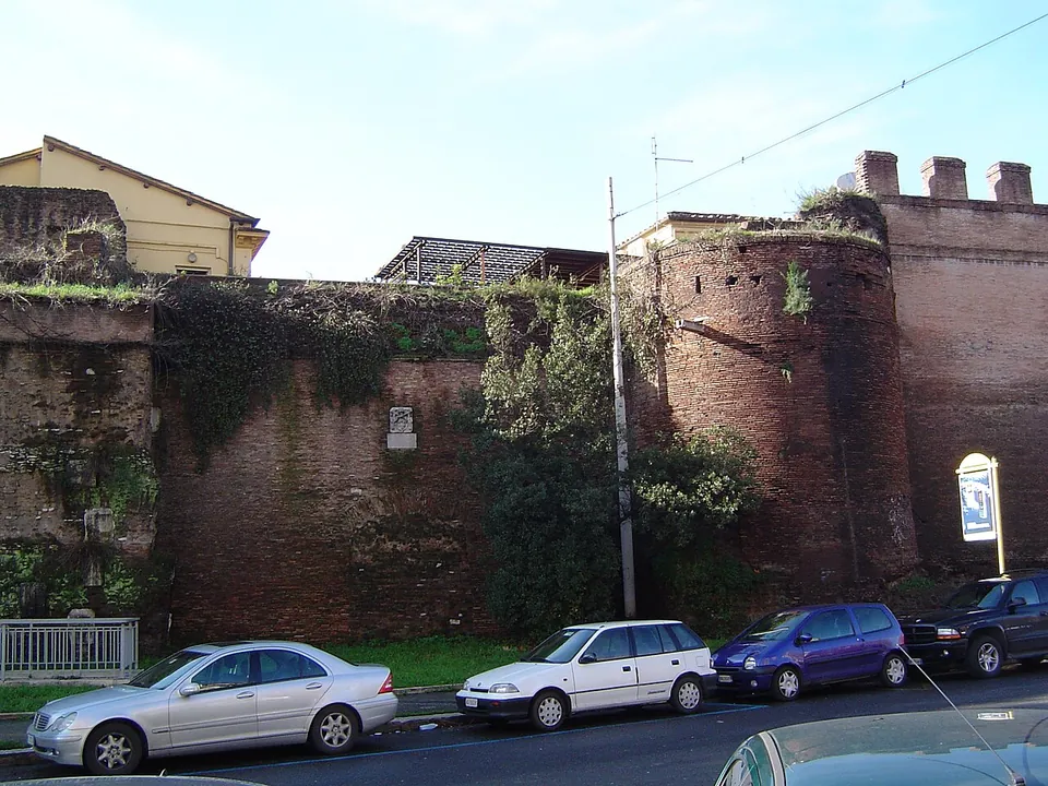 A stretch of high brick city wall along a street of parked cars, with a round brick tower at the right and a small white plaque set in the wall.