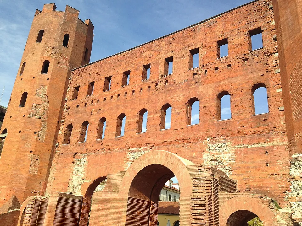 A close view up the brick face of the gate under a blue sky, a large round arch at the bottom, a row of arched windows above it and a row of square openings at the top, beside one of the many-sided towers.