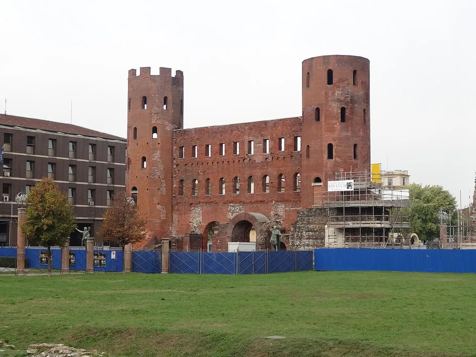 The Porta Palatina seen across a wide lawn, a long brick front pierced by two rows of windows between two tall many-sided towers, one of them crowned with battlements; scaffolding and a blue hoarding stand along its foot.