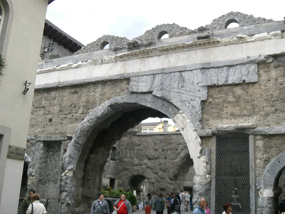 Close view of a stone arch whose rim and the band above it are faced with large pale grey marble slabs, with a ruined wall and small arched openings on top and visitors passing underneath.