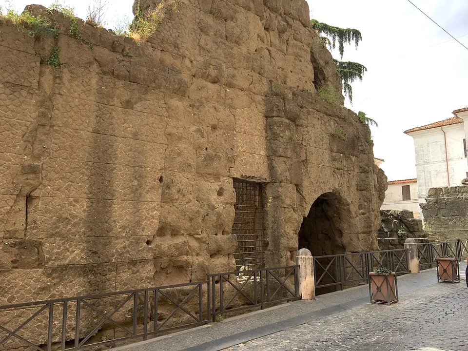 A massive wall of large, weathered tufa blocks rising above a cobbled street behind an iron railing, with a tall round-headed passage opening through it at the far end and a modern house beyond.