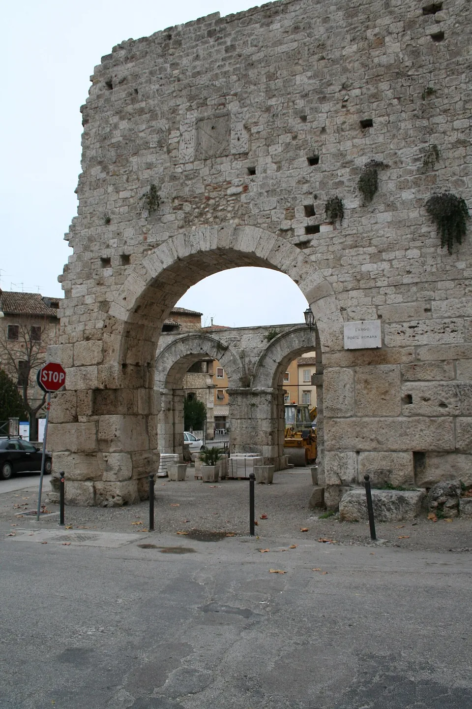 A tall wall of weathered squared stone blocks pierced by one round arch, with a street-name plaque reading Largo Porta Romana; through the arch, a few metres beyond, stands a lower double arch on stone piers, with paving stacked in front of it and houses behind.