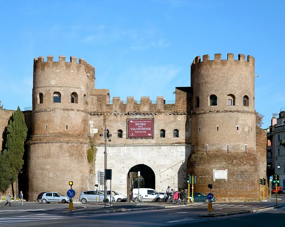 A gateway of two tall round crenellated brick towers joined by a stone-faced wall with a single arched passage, a red banner over the arch, cars parked at its foot.