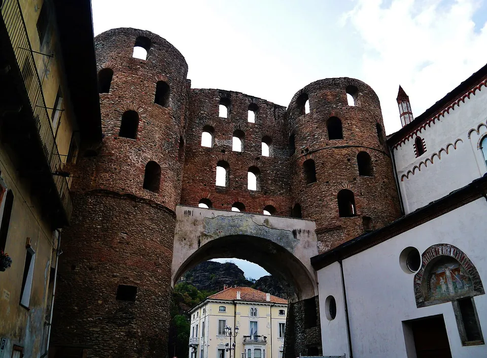 Looking up at the two round brick and stone towers and the wall between them, each pierced by staggered rows of arched openings, above a plain stone arch spanning the street; a white church wall with a painted lunette stands at the right.