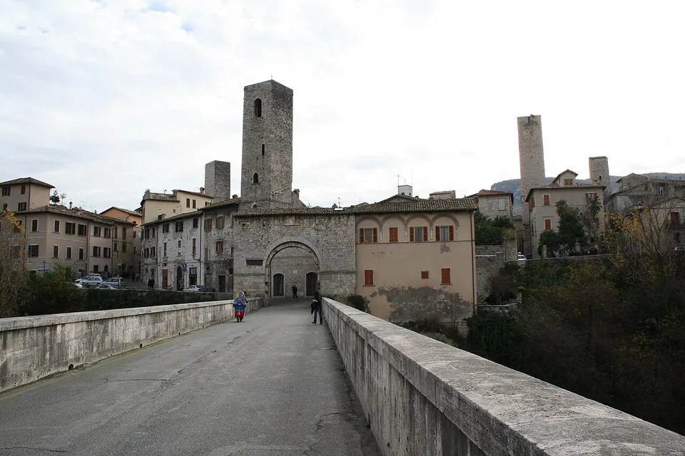 A road runs between low stone parapets across the bridge towards an arched stone gateway, with a tall square tower and houses rising behind it and a few people walking.