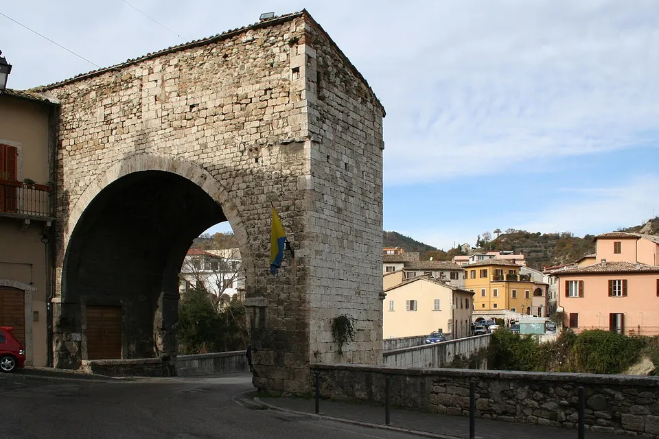 A tall block-shaped gateway of squared grey stone with one large round arch, a flag hanging from its side, standing at the end of a stone parapet with houses on the hillside behind.