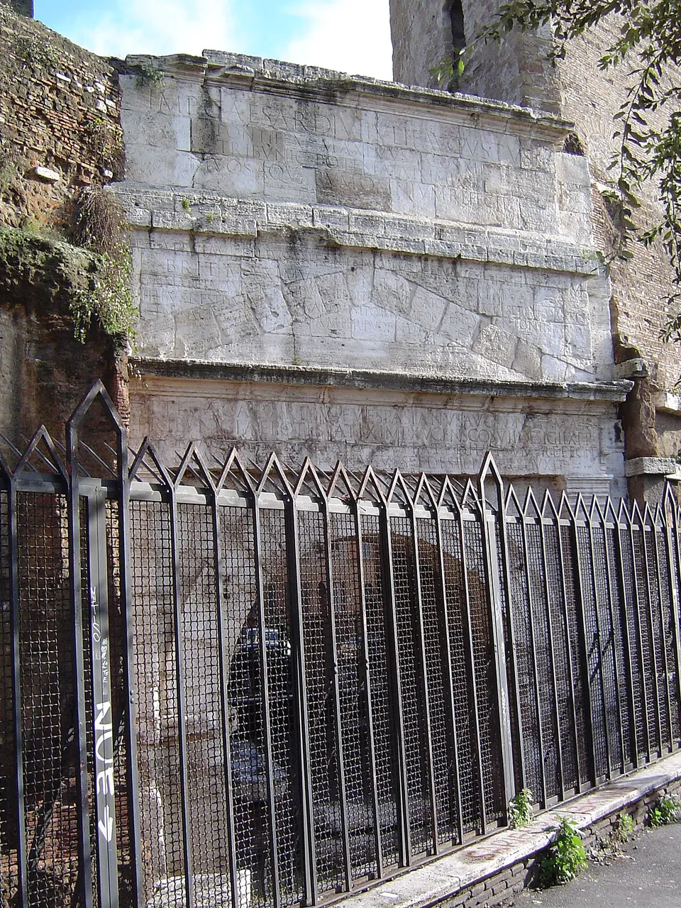 The upper part of an arch of large travertine blocks, carved with three bands of Latin capitals, the top line beginning "IMP CAESAR DIVI IVLI F", seen above a tall railing of black metal.