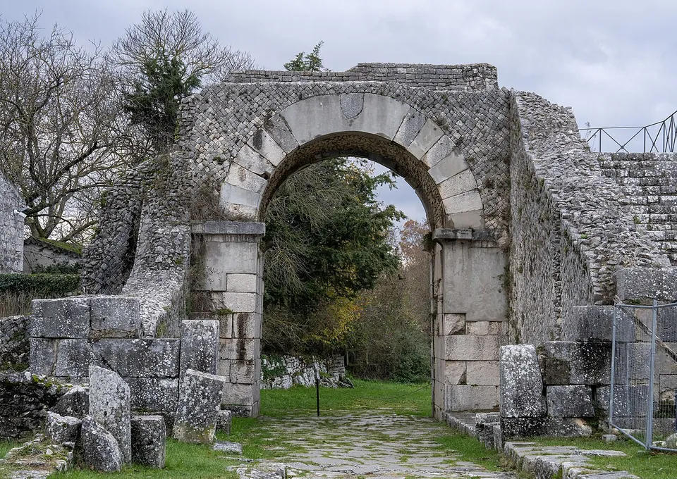 A round arch of large dressed stone blocks on square stone piers, set in a high wall faced with small stones laid in a diagonal net pattern, with fallen blocks in the grass on either side and a paved track running through the opening towards trees.