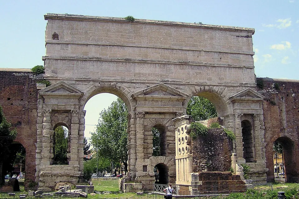 A massive double archway of rough-faced pale stone blocks under a tall plain attic, each pier framed by columns and a small pediment around a window, with brick wall continuing at both sides and a square block pierced by round openings standing in front of the gate.