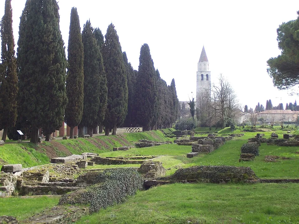 Low excavated stone walls and terraces in mown grass, running beside a tall row of dark cypresses, with a white bell tower and the roofs of the town in the distance.