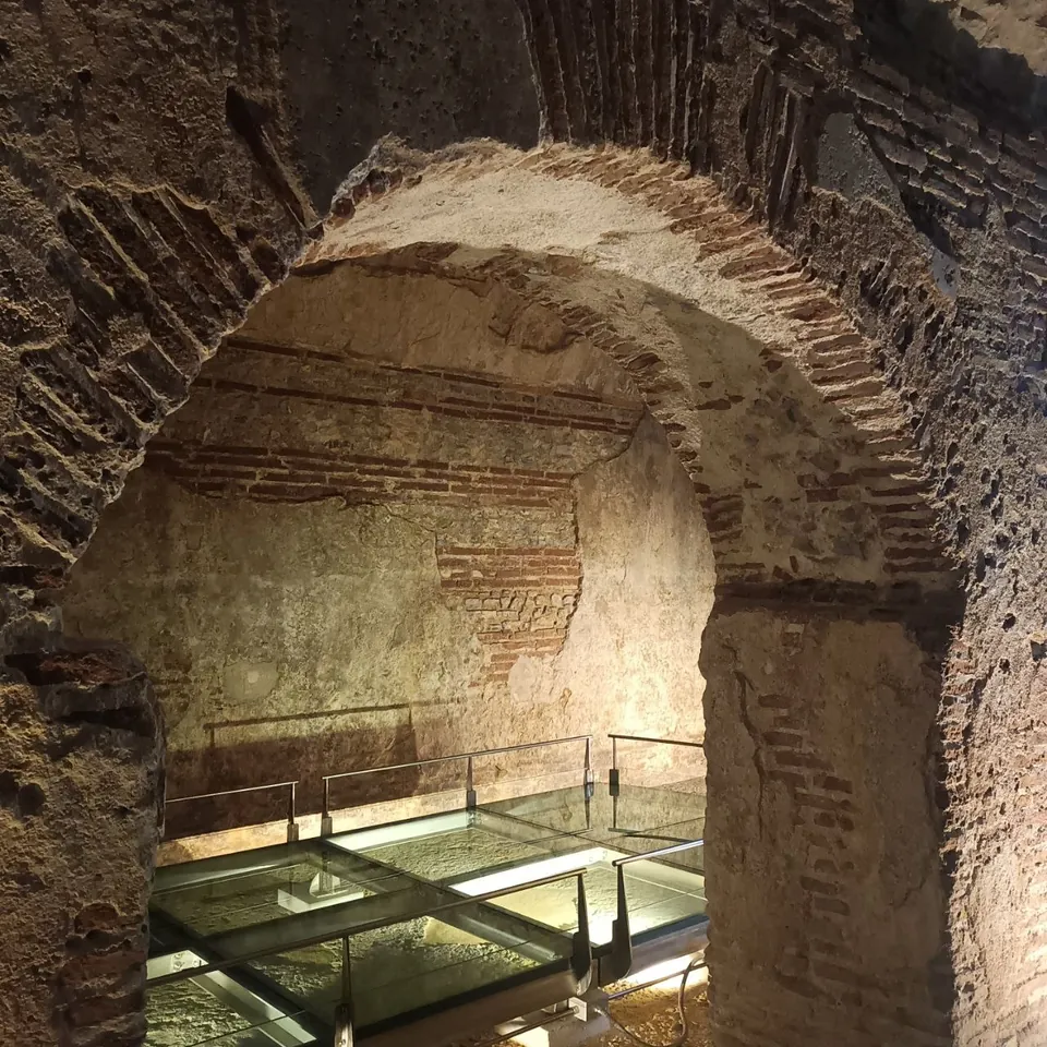 A lit interior of rough stone banded with brick, seen through a broad round arch, with a walkway of glass panels and a steel railing running along the floor.
