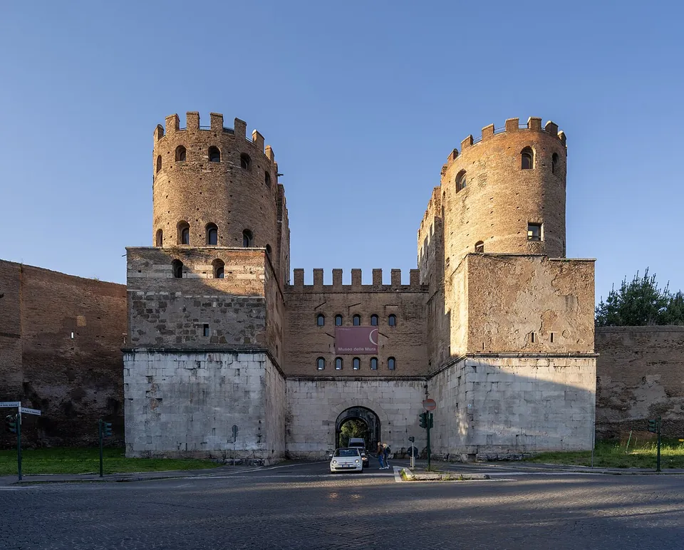 A gatehouse of two tall round crenellated brick towers rising from square bases faced in white stone, joined by a crenellated central block with rows of small arched windows over a single arched passage; the brick wall continues on either side.