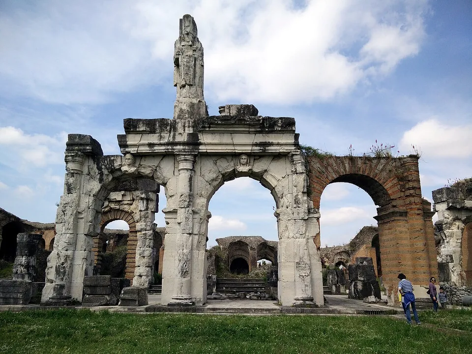 A tall arched opening in pale limestone, framed by carved half-columns and a cornice, with a broken stone pillar rising above it and a brick arch beside it, a lawn in front and two visitors at the right.