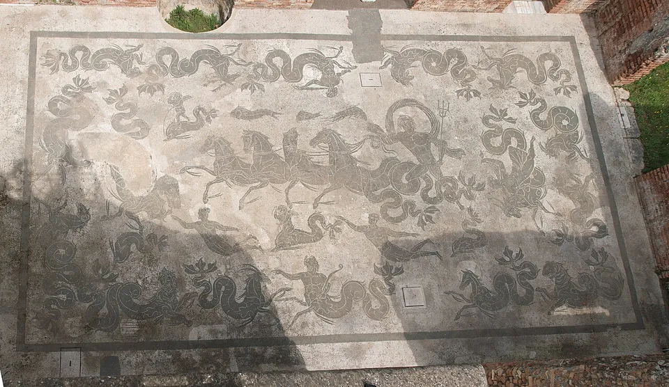 A large black-and-white floor mosaic, seen from above, of a figure driving a team of sea horses, ringed by swimming figures and coiling sea creatures.