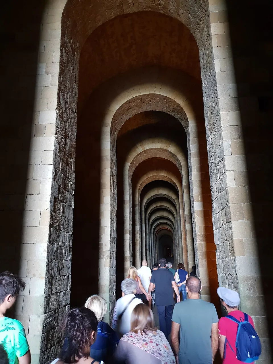 A group of visitors walking away from the camera down a long tunnel lined with a succession of tall masonry arches, lit from below.