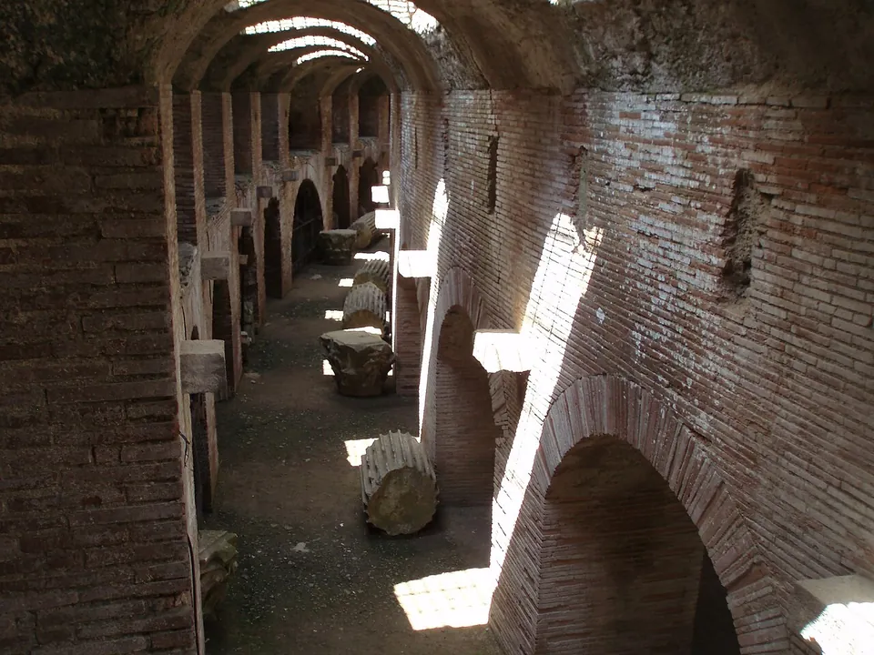 A long corridor with a brick barrel vault, lit by bright shafts of sunlight falling through openings above, with fragments of stone column shafts lying along the floor.