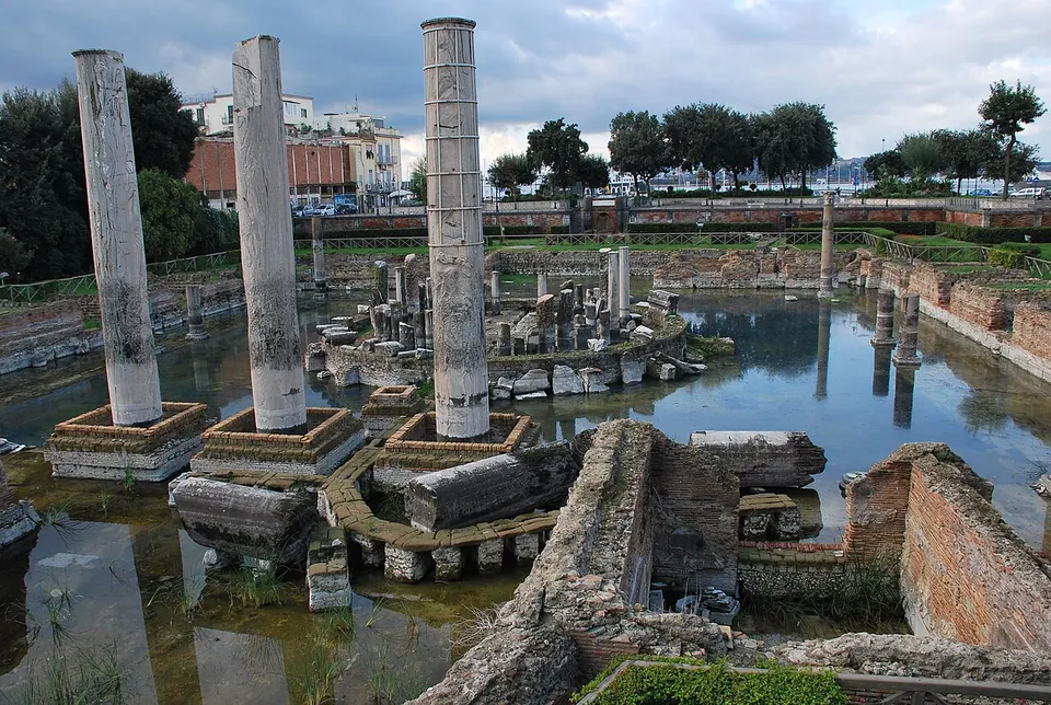 Three tall, plain marble columns standing in shallow water among the brick and stone foundations of a square courtyard, with a ring of broken column bases at its centre and trees and buildings behind.