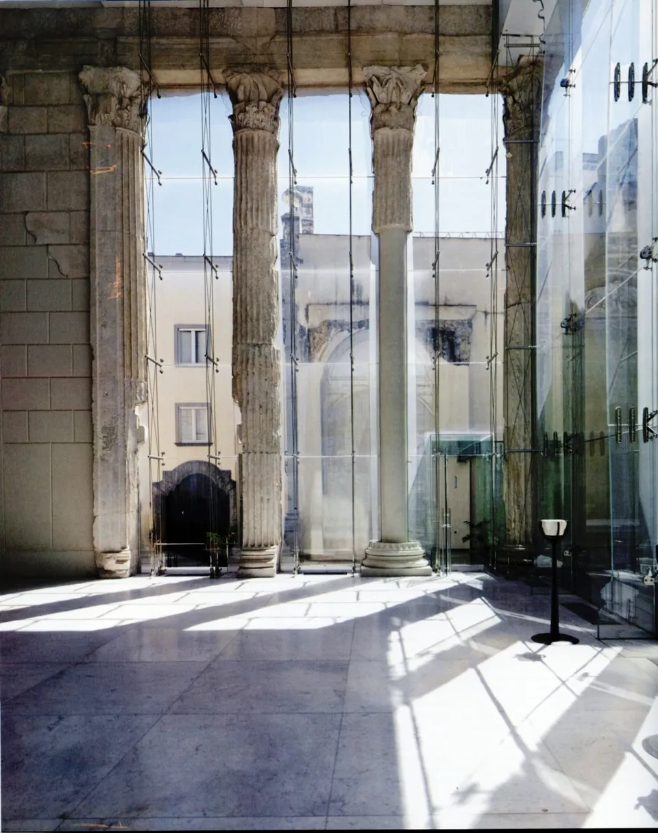 Inside a pale stone hall, fluted marble columns with carved leafy capitals stand in a row against a wall of glass panels, with sunlight on a polished stone floor.