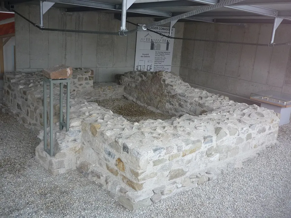 Low walls of pale stone, a few courses high, enclosing a rectangular space on gravel under a roof of steel girders, with a display panel against the back wall.