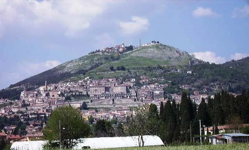 A town of pale houses climbing a green hillside under a cloudy sky, with a large building on broad terraces at its centre and a smaller cluster of buildings on the summit above.