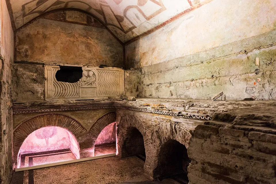An underground vaulted room with painted walls; a fluted marble sarcophagus with a round portrait panel sits in a wall niche above a row of small brick arches lit from below.