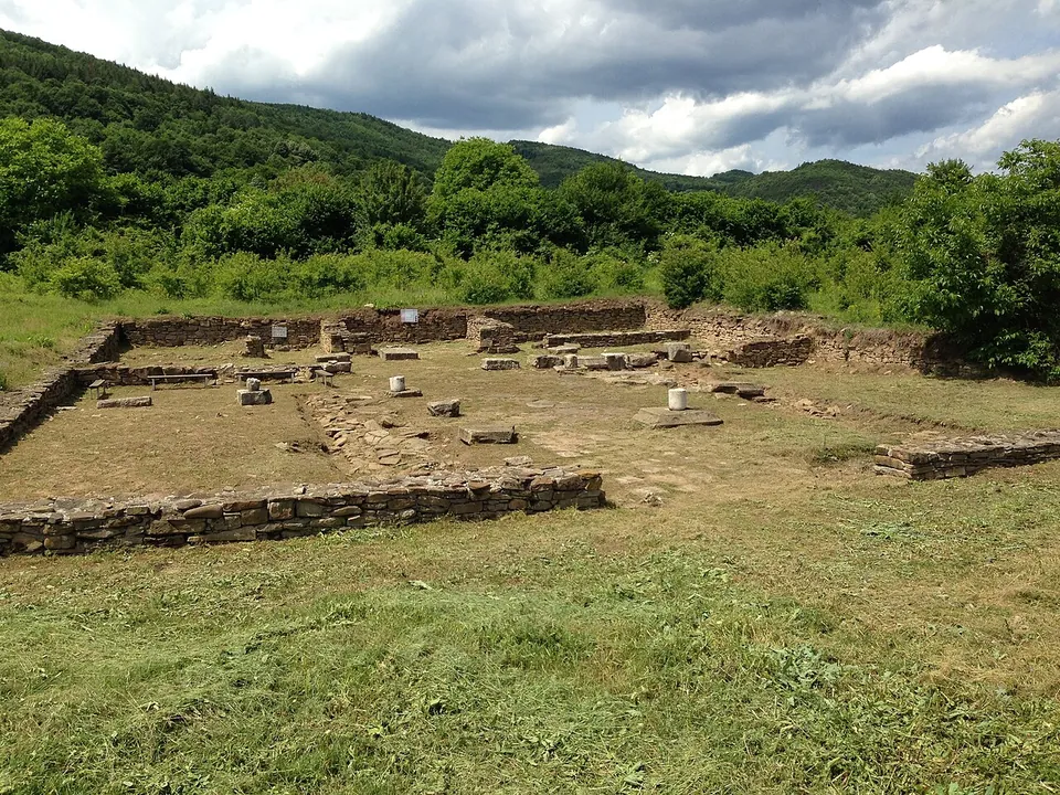 The low stone foundations of a large rectangular building laid out on mown grass, with column bases and blocks inside it and forested hills beyond.
