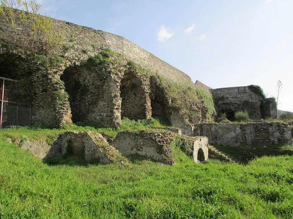 A row of tall concrete vaulted chambers opening onto a grassy terrace, the long retaining wall of a raised platform running behind them.