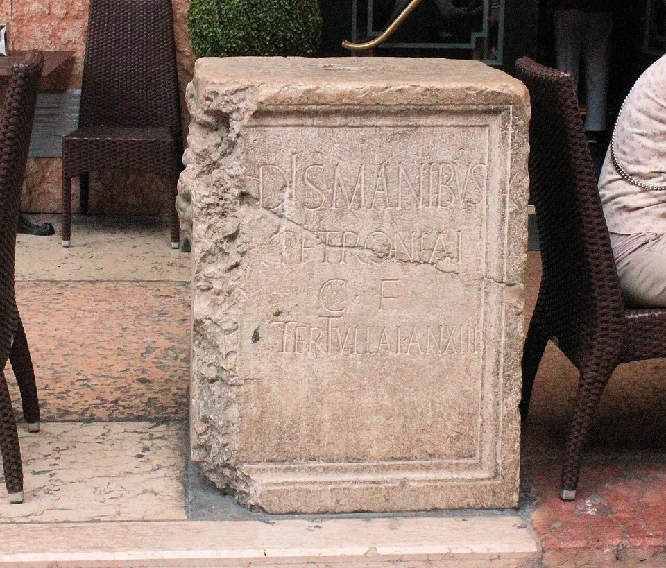 A squat block of stone with a moulded frame on its face, carved in Latin capitals DIS MANIBUS PETRONIAE C F TERTULLAE AN XIII, standing on the pavement between café chairs.