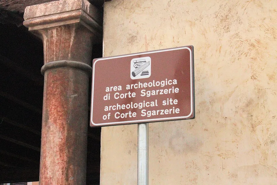 A brown sign reading "area archeologica di Corte Sgarzerie / archeological site of Corte Sgarzerie" on a post beside a red stone column of the loggia.