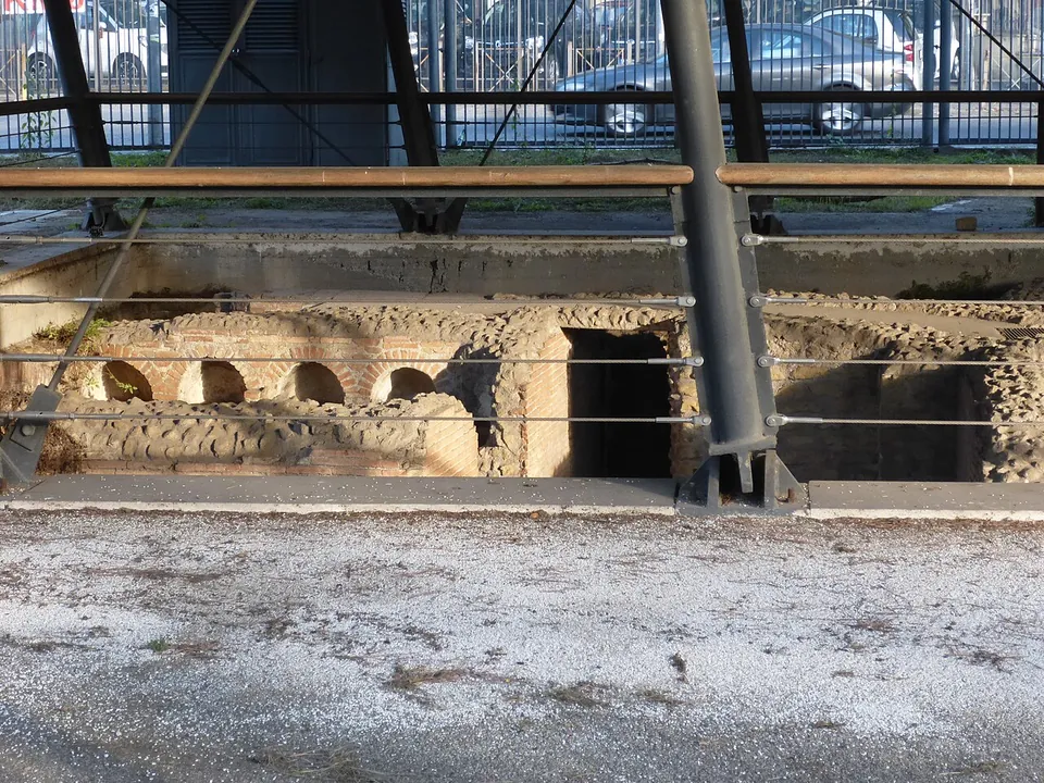 Low brick and stone tomb walls with small arched niches, sunk below ground level and seen through a metal railing, with parked cars beyond.