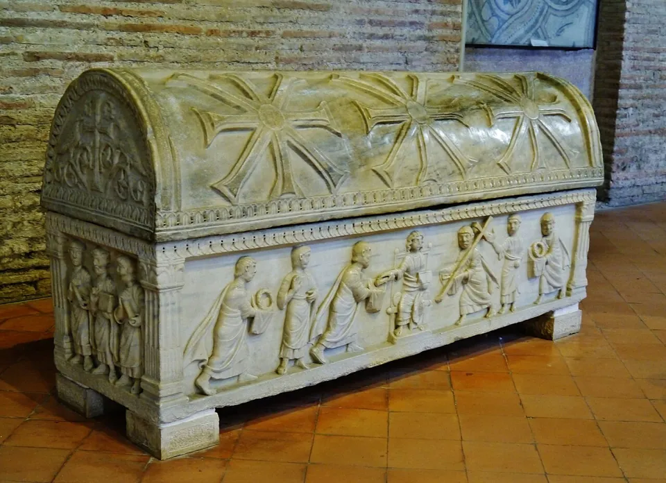 A white marble sarcophagus with a curved lid carved with large crosses; along its side a relief of robed figures walking towards a central figure, standing on a terracotta floor against a brick wall.