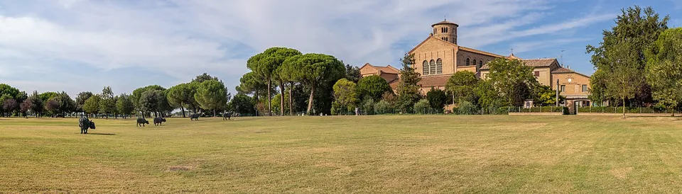 A wide mown meadow with umbrella pines and a few dark animal statues on the grass; at the right edge a long brick church with a tall round bell tower.