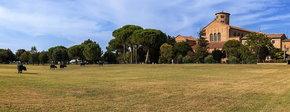 A broad mown meadow under a blue sky with umbrella pines, and beyond it a long brick church with a round bell tower.