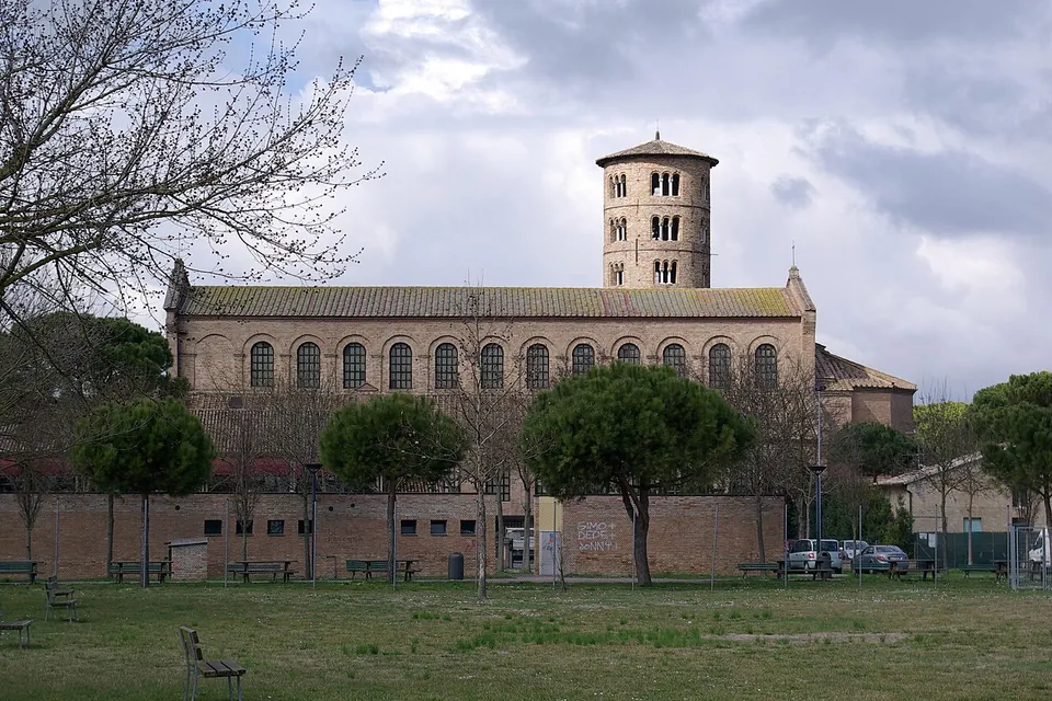 A long brick church with a row of tall round-arched windows along its upper wall and a tall cylindrical bell tower behind, seen across a lawn with young pines, a brick boundary wall and parked cars.