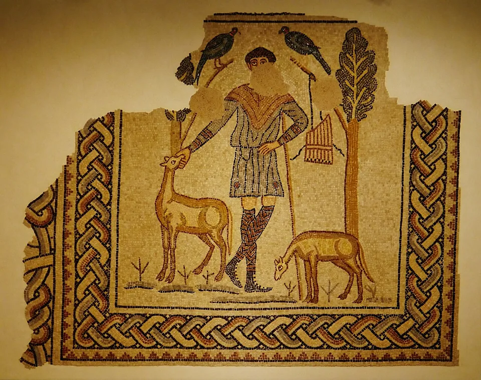 A mosaic panel mounted on a wall. A young man in a short striped tunic and patterned leggings, holding a staff, touches the head of a deer; a second deer grazes at the right, two birds perch on branches behind him and a set of pan pipes hangs from a tree at his side. The lower half of his face is lost. A broad plaited border runs round three sides.