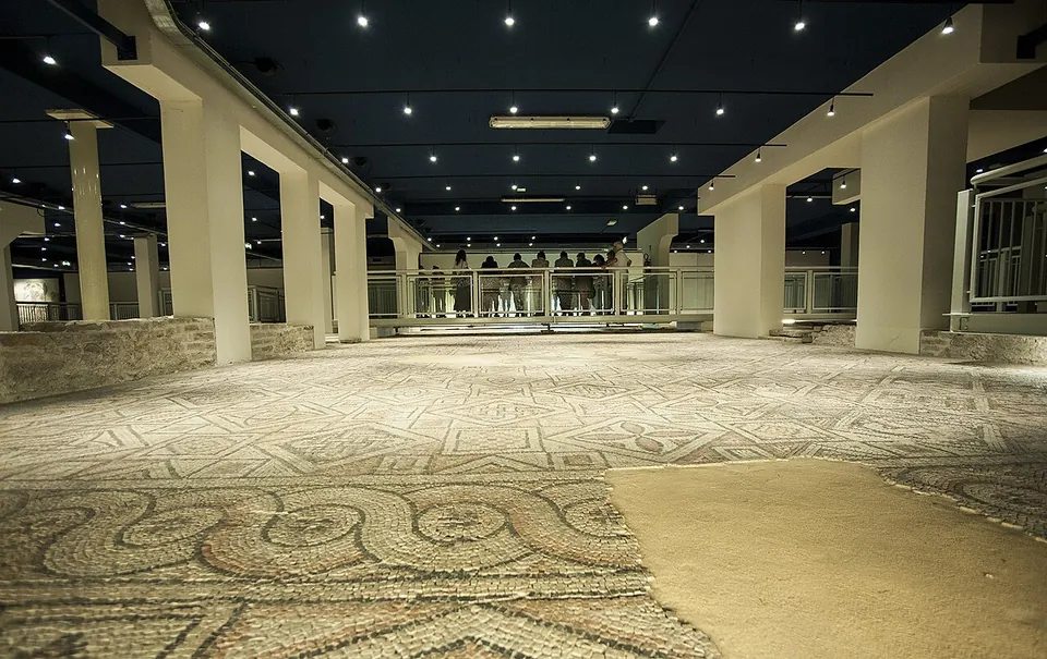 Mosaic floors in grey, white and ochre stretching away under a dark ceiling with spotlights, between rows of white pillars, with a railed walkway at the far end where visitors stand.