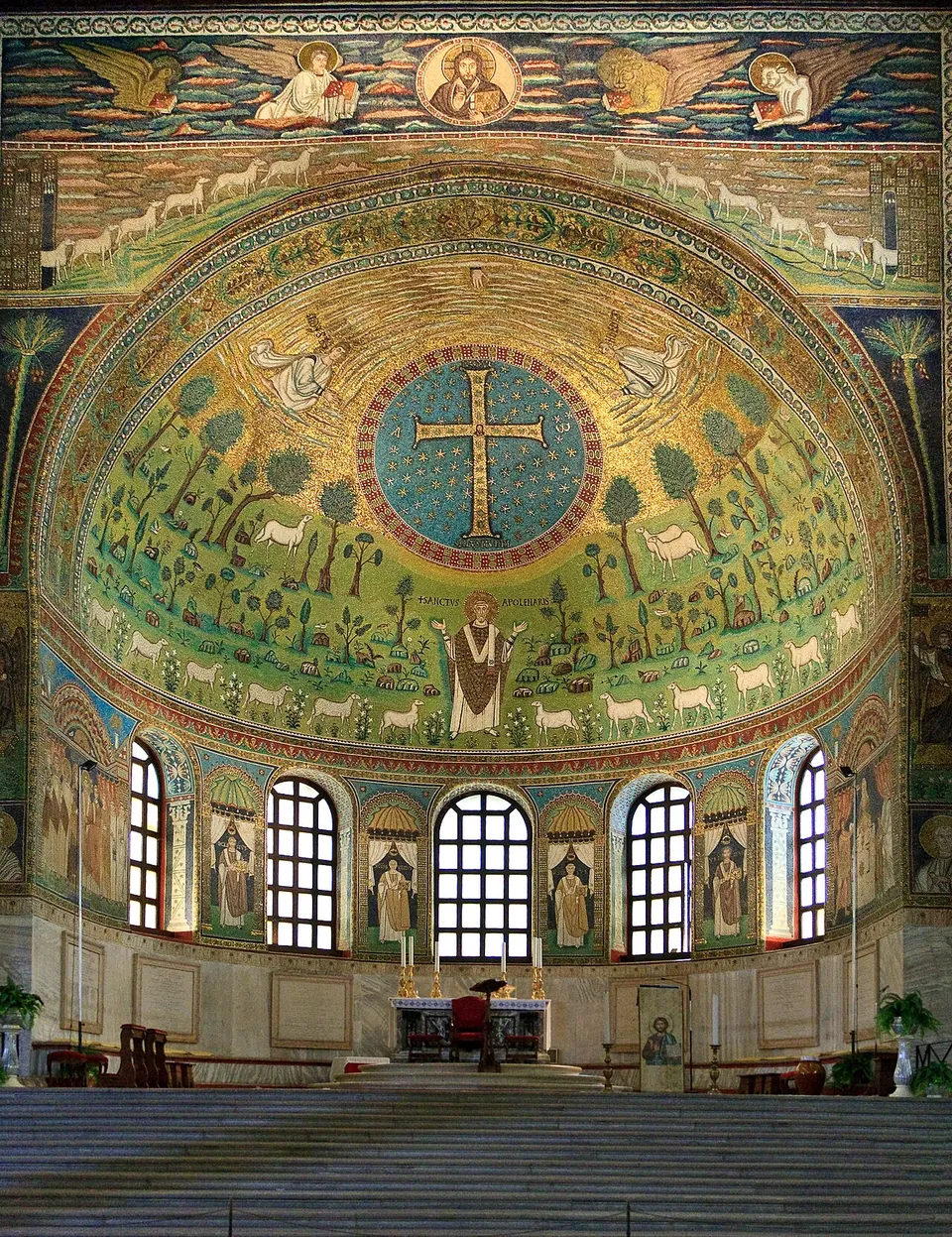 A half-dome of mosaic above three arched windows. In a ring of stars on blue at the top is a large jewelled cross; below it a green landscape of trees, rocks and birds, with a robed figure standing in the middle with raised hands between rows of white sheep. Above the arch, Christ in a medallion between winged figures.