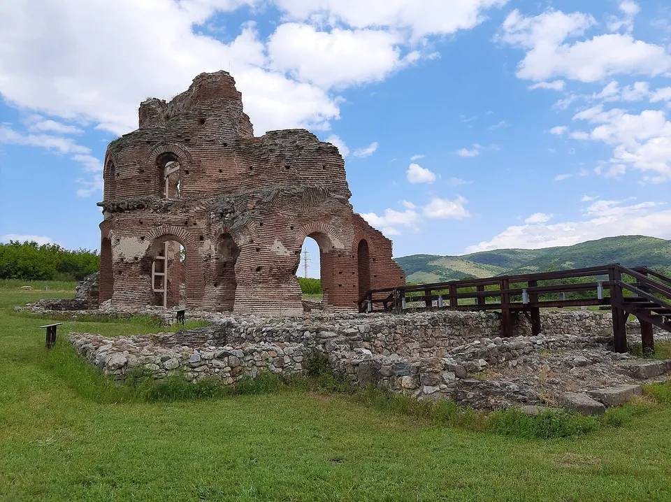 The tall ruined shell of a red-brick church with arched openings and the stump of a dome, standing on a lawn behind low stone foundations, with a wooden walkway on the right and green hills under a cloudy sky.