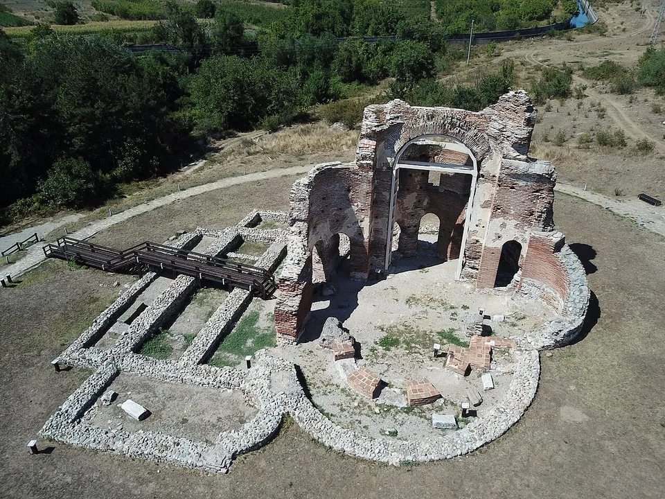 An aerial view of the ruined brick church, its tall walls and arches braced by a metal frame, with the low outlines of the narthexes and side rooms laid out in stone on the ground and a wooden walkway beside them.