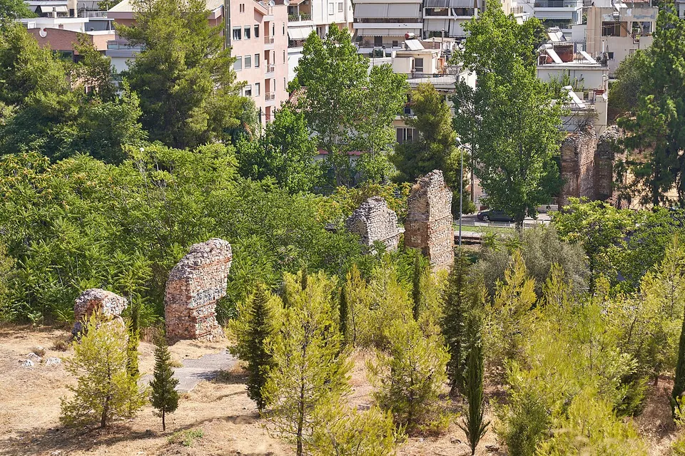 A line of tall ruined piers of stone and brick standing among pines and shrubs on a slope, with apartment blocks behind.